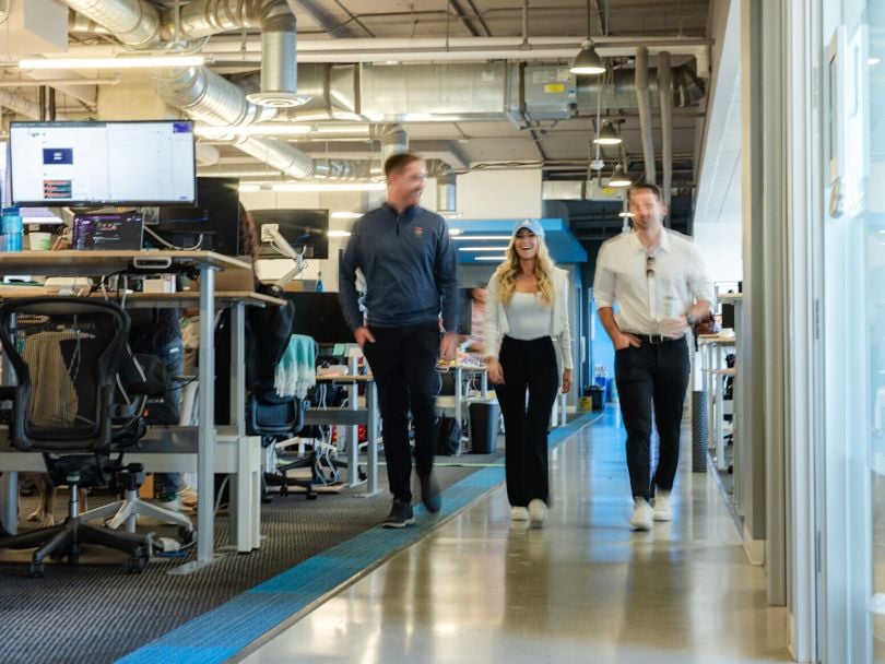 Three Crexi team members chat together while walking past rows of desks in the company’s office